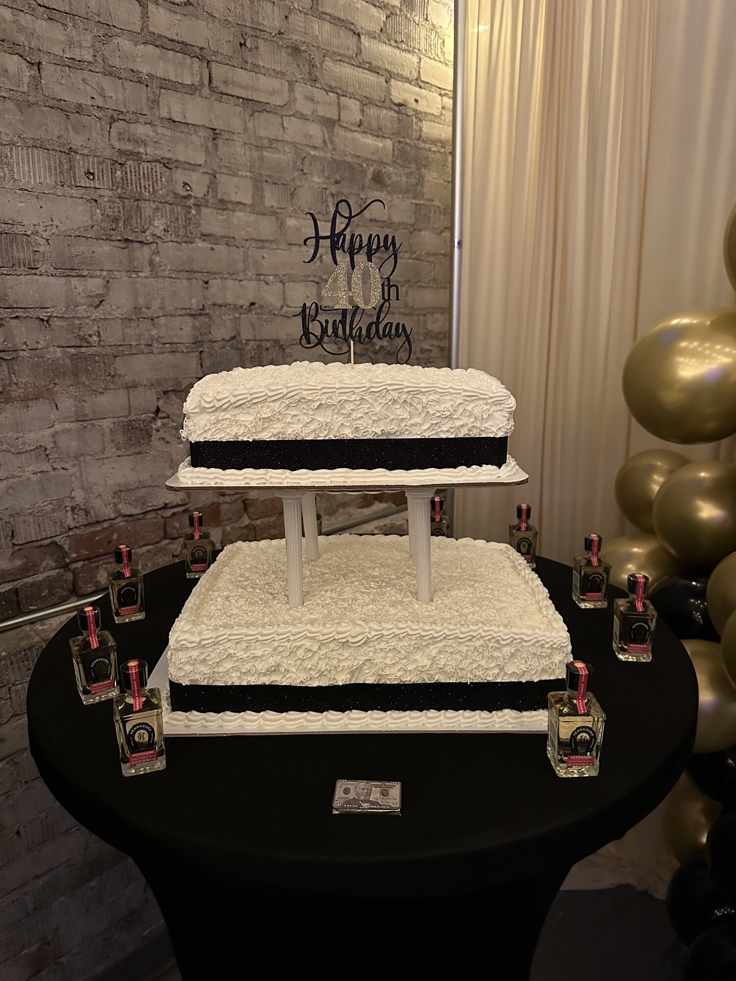 Two-tier Happy 40th Birthday cake on a black cocktail table against exposed brick