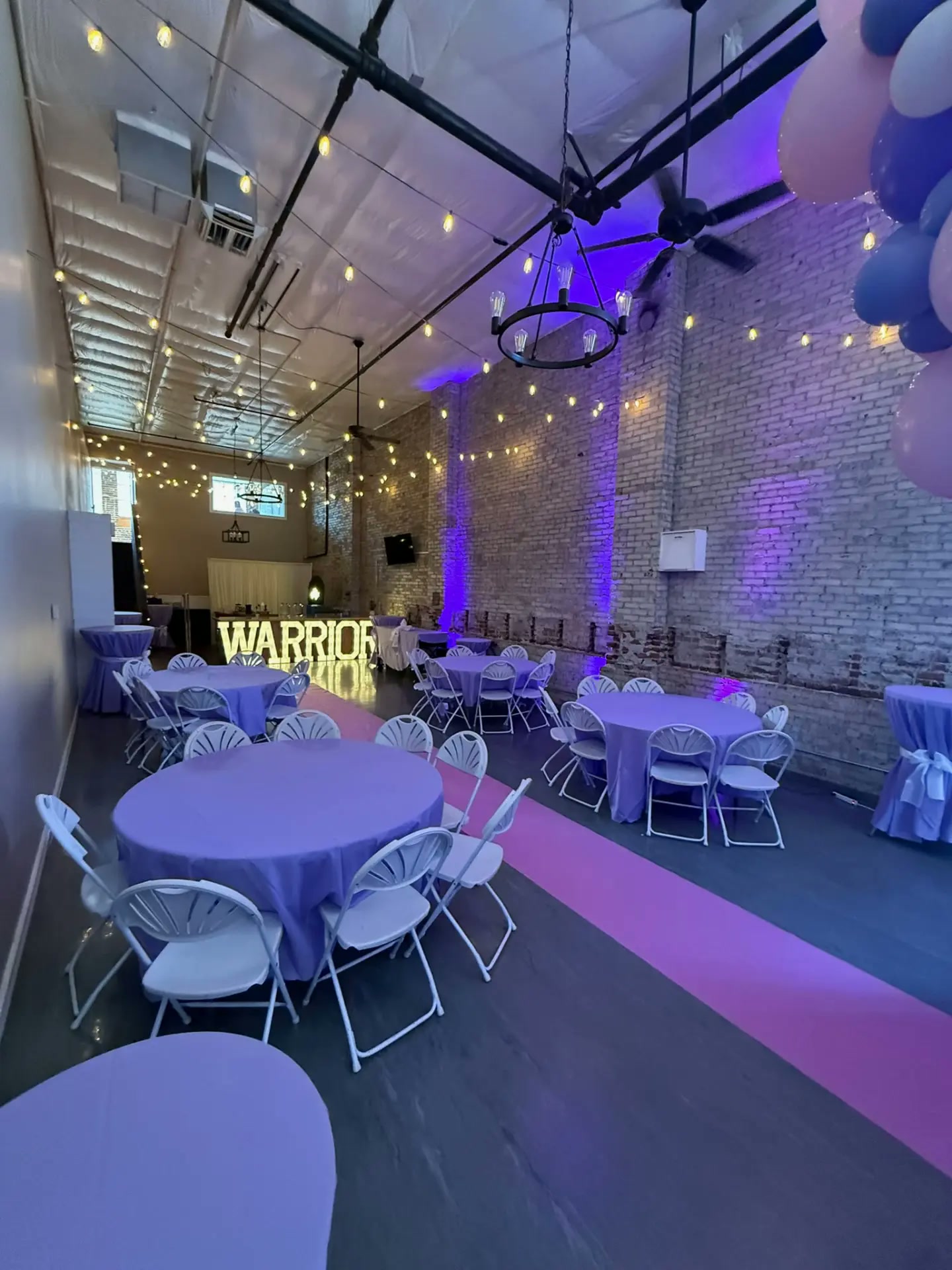 Wide view of a purple birthday party with WARRIOR marquee letters, string lights and balloon garland at Host Wenatchee