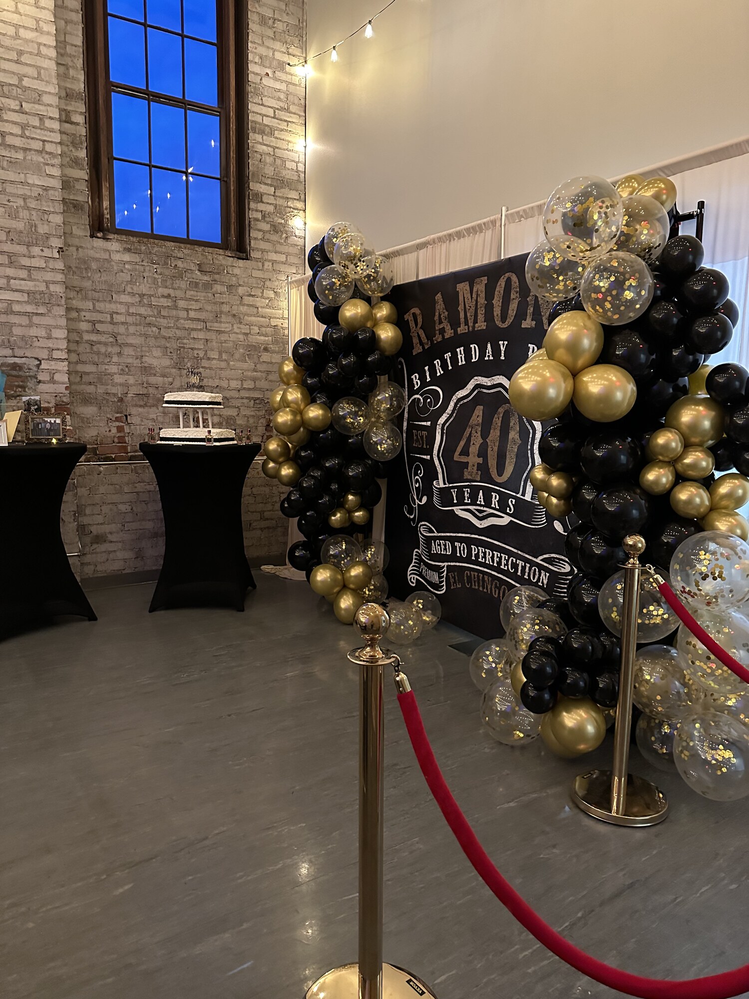 Red velvet rope entrance beside a black and gold balloon backdrop at a milestone birthday party