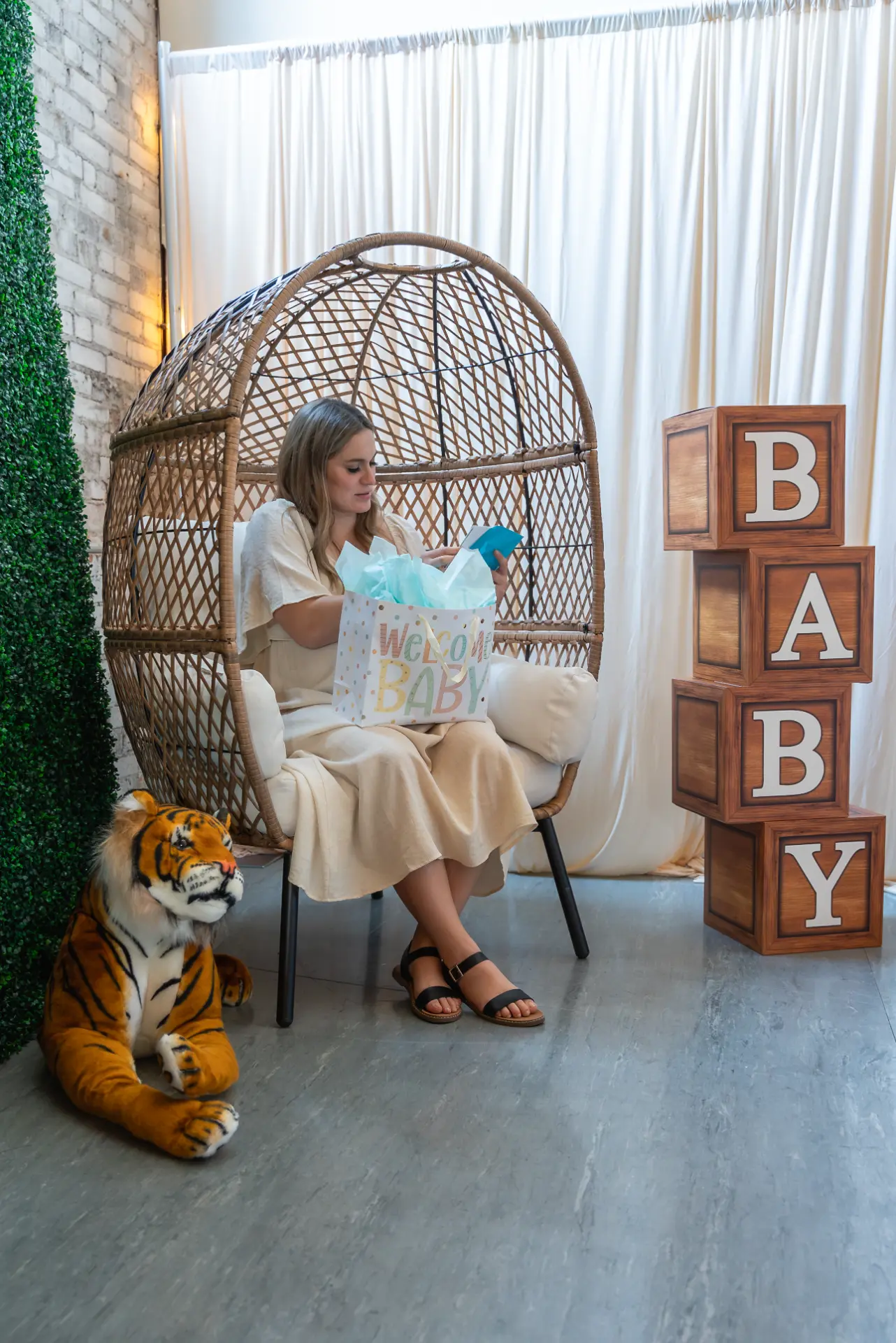 Mom-to-be in a wicker egg chair beside wooden BABY blocks, a greenery wall and a plush tiger at a jungle baby shower in Wenatchee