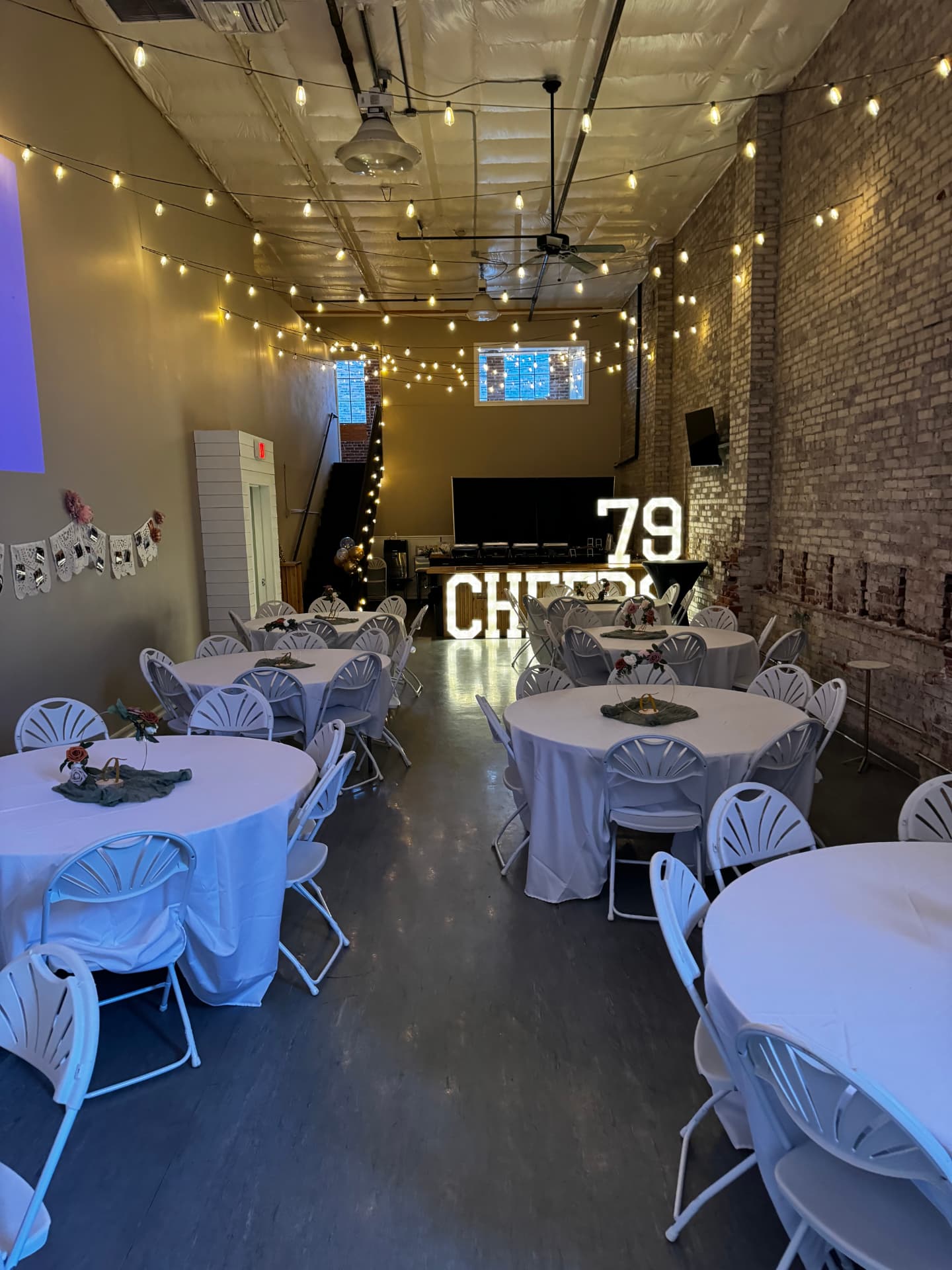 White-linen round tables with floral centerpieces and glowing CHEERS marquee letters at a holiday celebration in downtown Wenatchee