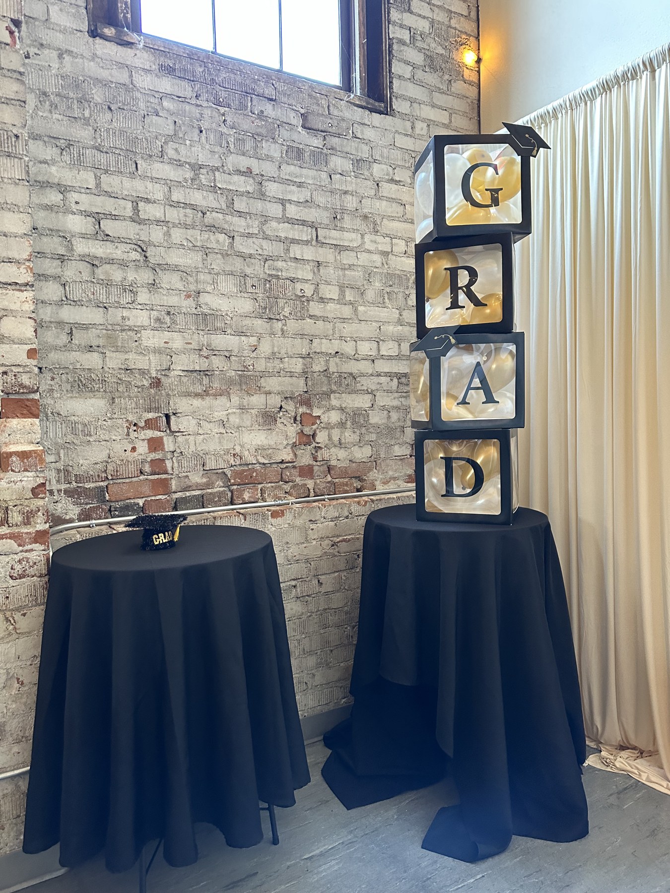 Black and gold GRAD balloon boxes beside cocktail tables against the exposed brick wall at Host Wenatchee