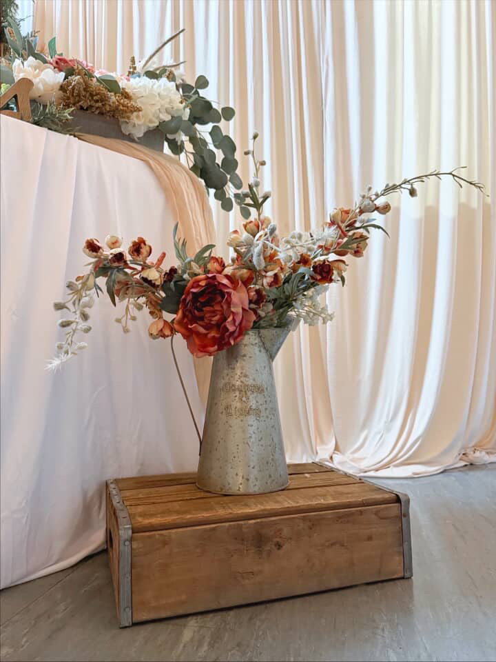 Galvanized pitcher of roses and eucalyptus on a wood crate beside a draped wedding table