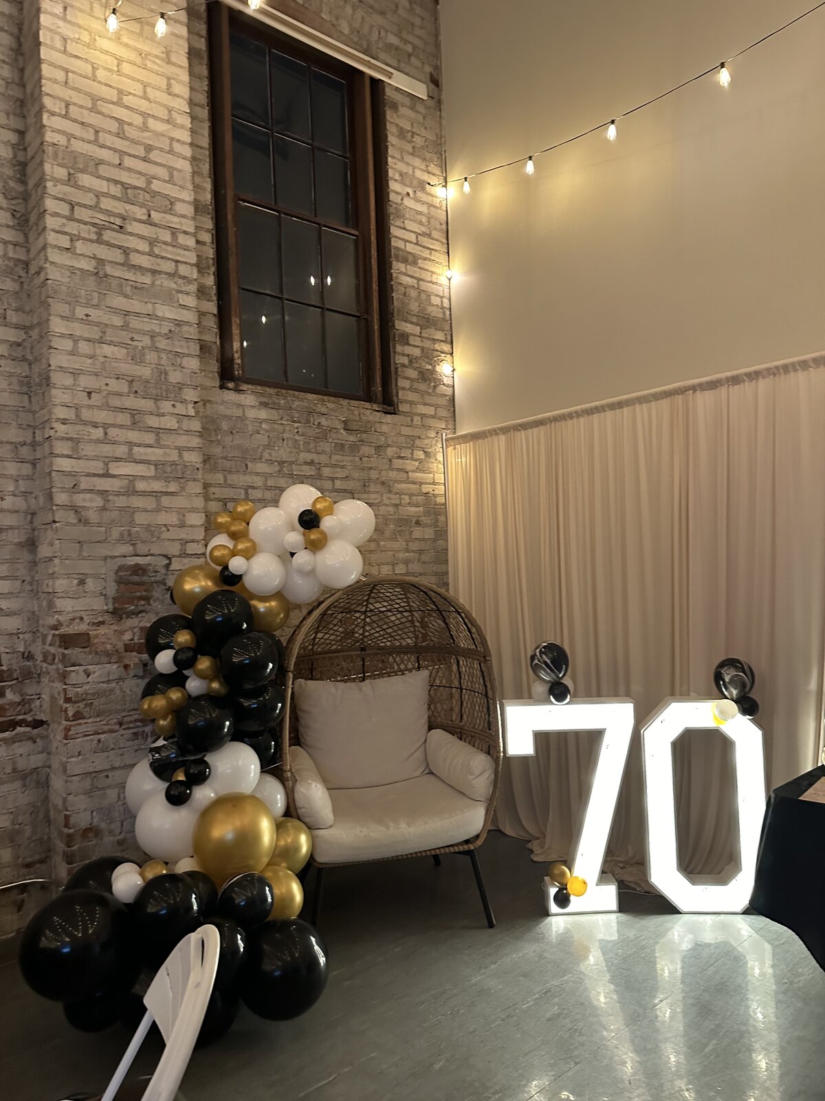 Light-up 70 marquee numbers, black and gold balloon garland and a wicker egg chair photo corner at a 70th birthday party in Wenatchee