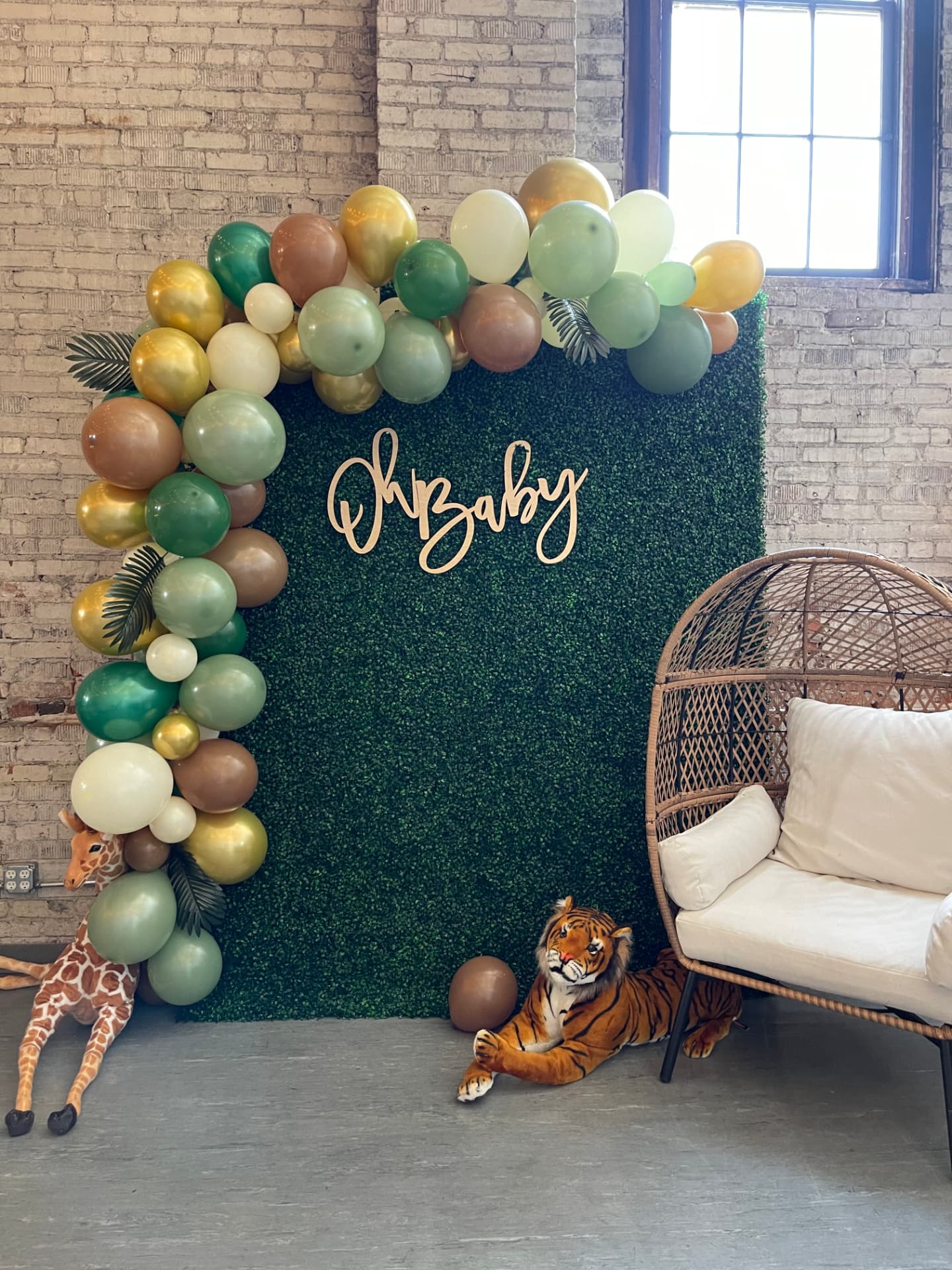 Jungle-theme Oh Baby greenery backdrop with green and gold balloon arch, giraffe and wicker egg chair at Host Wenatchee